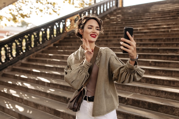 Cheerful woman in cool jacket and light jeans making selfie. Young woman with wavy hair shows peace sign outside.