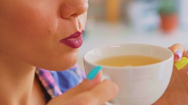 Cheerful woman drinking hot green tea in the morning. Close up of pretty lady sitting in the kitchen in the morning during breakfast time relaxing with tasty natural herbal tea from white teacup.
