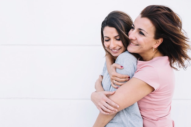 Cheerful woman embracing woman near white wall