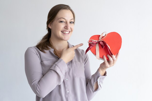 Cheerful woman holding heart shaped gift box and pointing at it