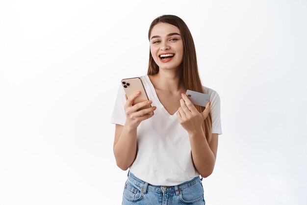 スマートフォンとクレジットカードを持っている陽気な女性白い背景の上に立っているモバイルアプリで買い物をしている間、電話で笑ってオンラインで非接触型決済をしている幸せな女の子
