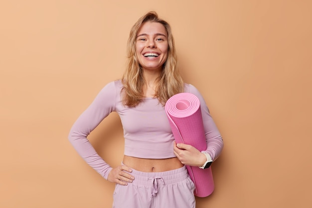Cheerful woman leads sporty lifestyle dressed in activewear hold rolled mat being in good mood prepares for training in gym smiles broadly isolated over brown background Regular workout concept