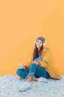 Free photo cheerful woman listening music on smart phone sitting on rug against yellow surface