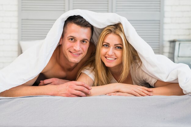 毛布の下でベッドの若い笑っている男の近くに横たわる朗らかな女性