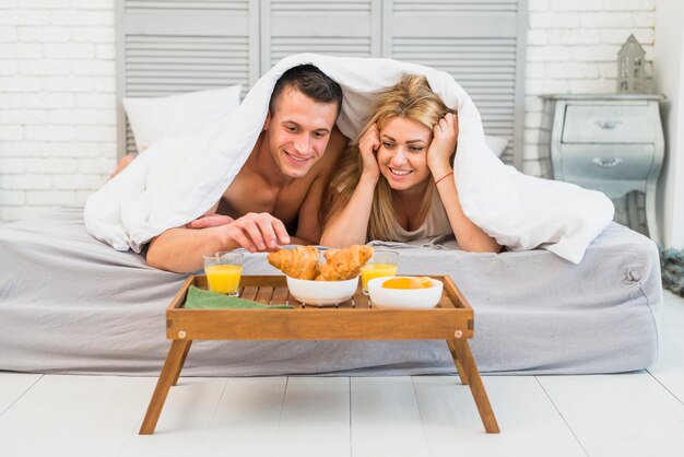 Donna allegra vicino al giovane a letto sotto coperta vicino al cibo sul tavolo della colazione