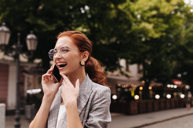 Cheerful woman in plaid outfit and eyeglasses happily talking on phone