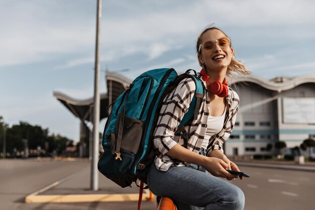 Cheerful woman in plaid shirt and denim pants smiles widely near airport Blonde girl in sunglasses holds backpack and phone Tourist in denim pants and white tee poses