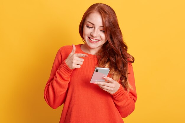 Cheerful woman pointing to her smart phone screen with index finger, looking at mobile phone with charming smile