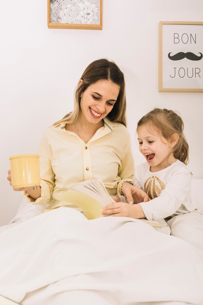 Cheerful woman reading book with daughter in bed