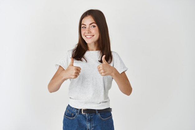 Cheerful woman saying yes, showing thumbs-up in approval, like and agree with you, standing over white wall.