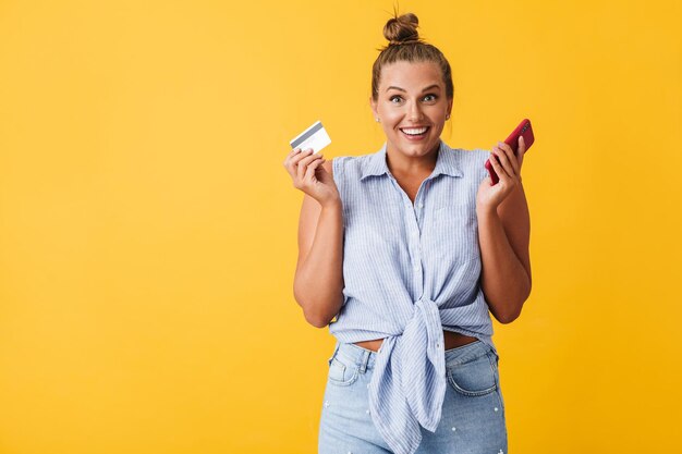 Cheerful woman in shirt joyfully looking in camera while holding credit card and cellphone in hands over yellow background