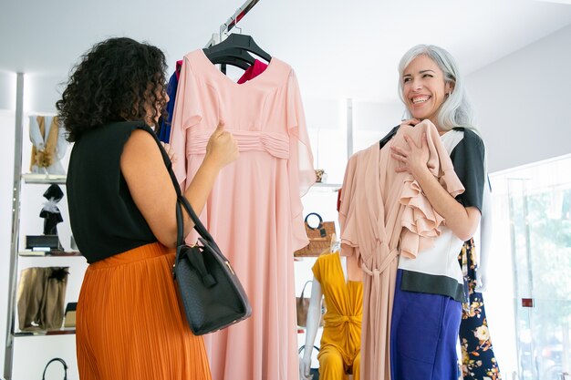 Cheerful woman showing chosen dress with hanger to her friend and laughing. Two ladies shopping in fashion store together. Consumerism or shopping concept