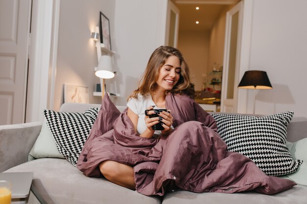 Cheerful woman sitting on couch with blanket and cushions and smiling
