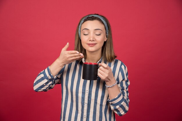 Cheerful woman smells aroma of coffee on red wall