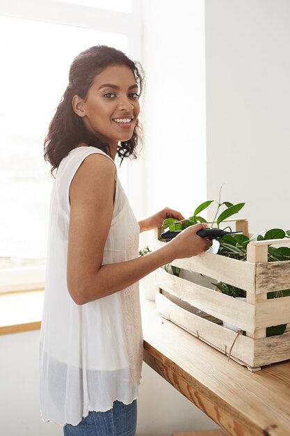 Cheerful woman smiling preparing to cut plant stems over white wall and window