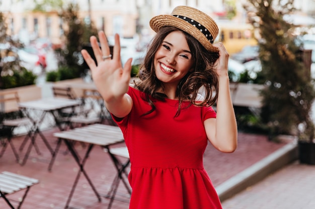 Cheerful woman in straw hat and red dress waving hand to camera. Blithesome cute girl expressing good emotions.