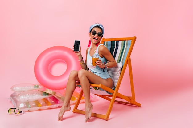 Free photo cheerful woman in sunglasses sitting on deck chair with gadget