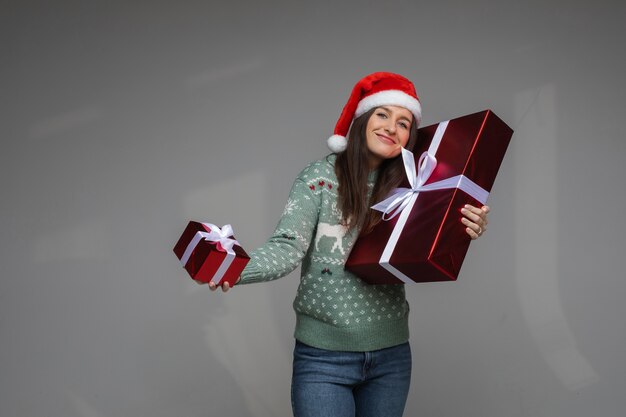 스웨터와 cristmas 모자를 쓴 쾌활한 여성이 크리스마스 선물을 들고 상자를 기뻐합니다