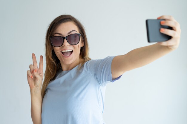 スマートフォンでselfie写真を撮っている明るい女性。勝利のサインを示す幸せな女性。