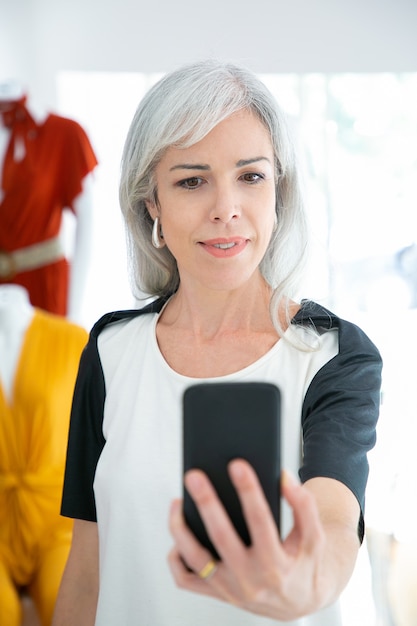 Cheerful woman taking selfie on smartphone while shopping in fashion store. Medium shot, front view. Boutique customer or communication concept