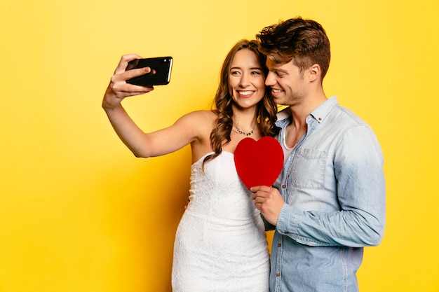 Cheerful woman taking selfie on smartphone with her attractive boyfriend holding red heart