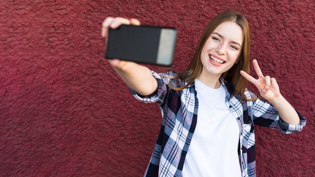 Cheerful woman taking selfie with peace sign on rough wall textured backdrop