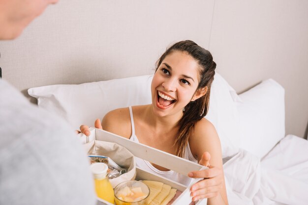 Cheerful woman taking tray with breakfast in bed