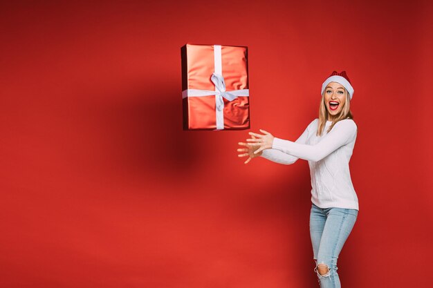 陽気な女性がクリスマスプレゼント、赤い背景で隔離の画像で大きな箱を投げる