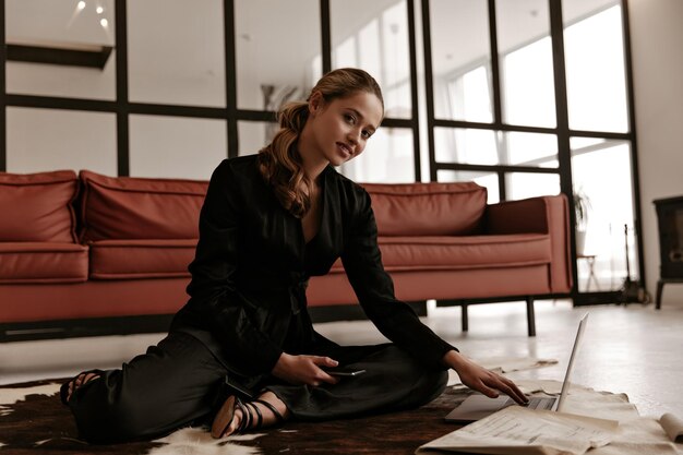 최신 유행의 검은 실크 수트를 입은 쾌활한 여성이 바닥에 깔린 카펫에 앉아 카메라를 쳐다보고 있습니다.