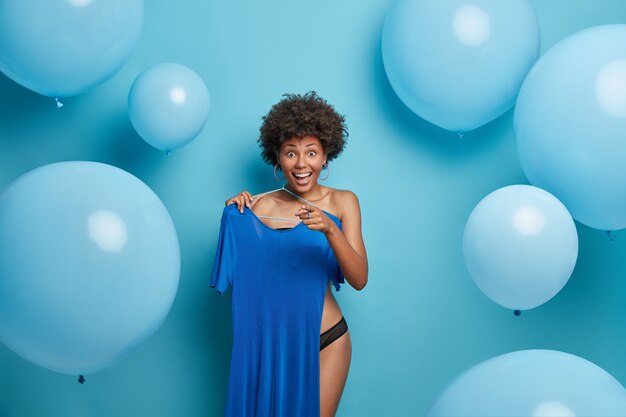 Cheerful woman in underwear, chooses best outfit, prepares for important meeting, holds blue elegant dress on hanger, points at you and smiles broadly, poses against inflated helium balloons