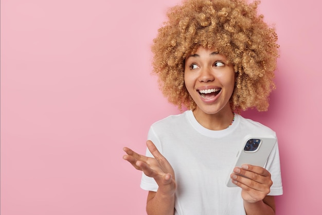 Cheerful woman uses mobile phone application communicates online laughs gladfully dressed in casual white t shirt isolated over pink background with empty space for your advertising content