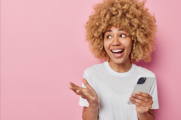 Cheerful woman uses mobile phone application communicates online laughs gladfully dressed in casual white t shirt isolated over pink background with empty space for your advertising content