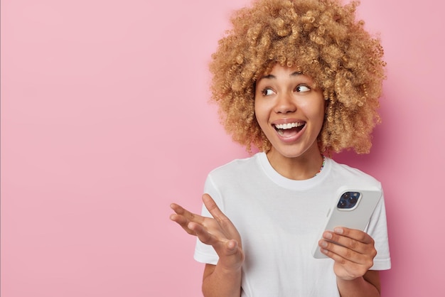 쾌활한 여성은 휴대 전화 응용 프로그램을 사용하여 광고 콘텐츠를 위한 빈 공간이 있는 분홍색 배경 위에 격리된 캐주얼한 흰색 티셔츠를 입고 기쁘게 온라인 웃음을 전달합니다.