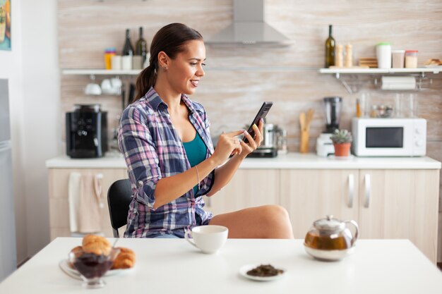 朝食と緑茶の間にキッチンでスマートフォンを使用して陽気な女性