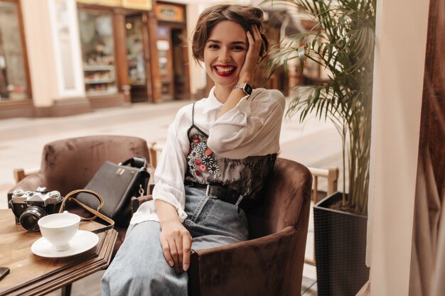 Cheerful woman with brunette hair and red lips laughing in restaurant. Optimistic woman in blouse and jeans poses in cafe.