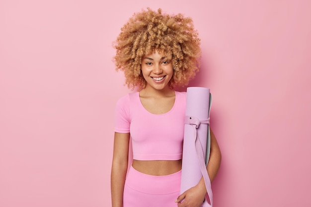 巻き毛の陽気な女性は、スポーティーな服を着て、ピンクの背景の上に隔離された自宅でフィットネストレーニングの準備ができてロールマットを保持します幸せなスポーティーな女性モデルは、ヨガの練習のためのカレムトを運びます