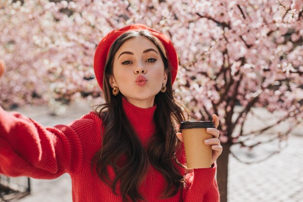 お茶を片手に元気な女性がキスをして自撮りします。咲く桜に対してコーヒーカップを保持している赤いセーターの女性の肖像画