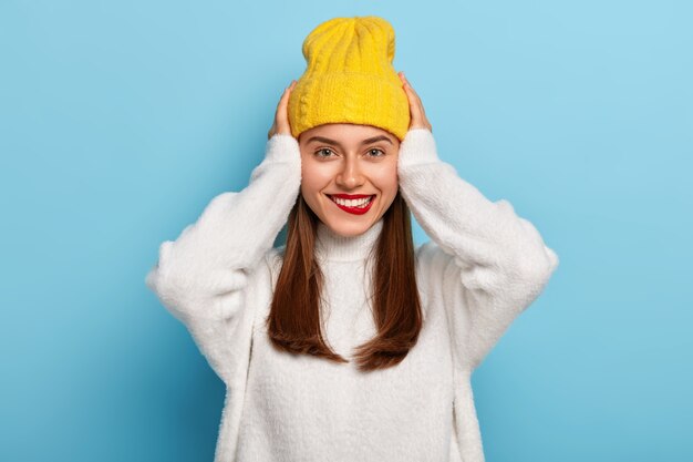 메이크업, 밝은 빨간색 립스틱을 가진 쾌활한 여자는 두 손을 머리에 유지하고 하얀 치아를 보여주고 모자와 캐주얼 스웨터를 입습니다.