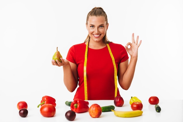 Cheerful woman with measuring tape on neck holding pear in hand and showing ok gesture while happily looking in camera with fruits and vegetables near over white background