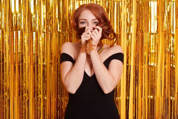 Cheerful woman with red hair wears black dress, poses over tinsel curtain, has fun at night club, covering her face with her hair, expressing positive emotions and flirt.