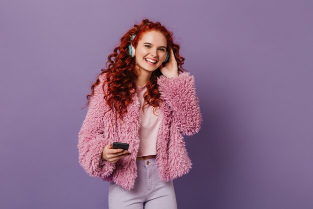 Cheerful woman with red locks dressed in pink eco-coat laughs while listening to song in headphones on lilac space.