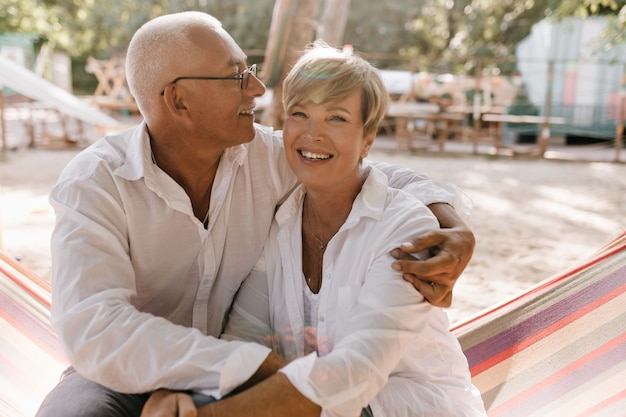 Free photo cheerful woman with short blonde hairstyle in white clothes sitting on hammock and hugging with smiling man in eyeglasses on beach.