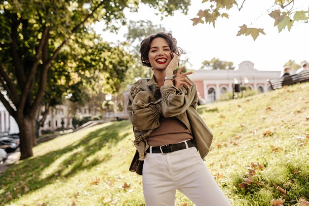 Cheerful woman with short curly hair in white pants with belt smiling outside. Trendy lady in denim olive jacket laughing outdoors.