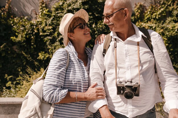 Cheerful woman with short hair and backpack in sunglasses and striped blouse looking at man with mustache in white shirt with camera outdoor.