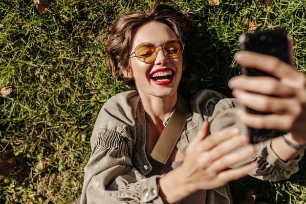 Cheerful woman with short hair in yellow glasses laughs and lies on grass outside. woman in denim jacket making selfie outdoors.