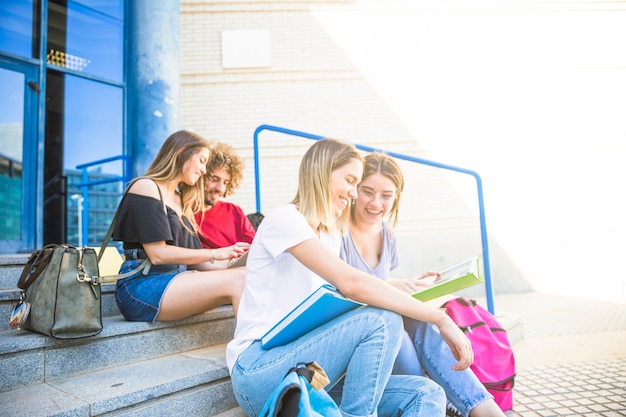 Cheerful women studying on university stairs near friends