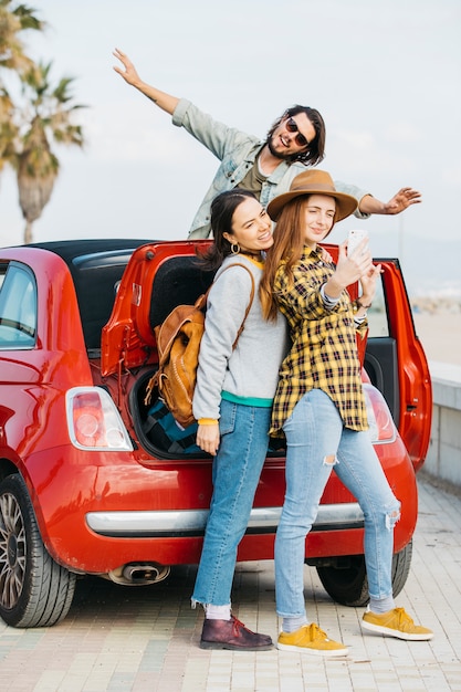 Cheerful women taking selfie on smartphone near car trunk and man leaning out from auto