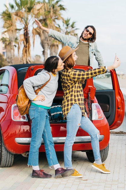 陽気な女性が車のトランクと自動車から傾いている人の近くのスマートフォンでselfieを取って