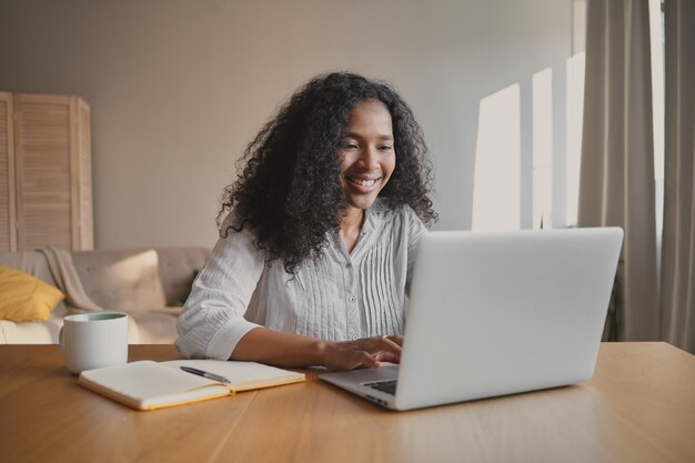 Cheerful young African American woman copywriter sitting in front of open laptop with mug and copybook on desk, feeling inspired, working on new motivation article. People, occupation and creativity