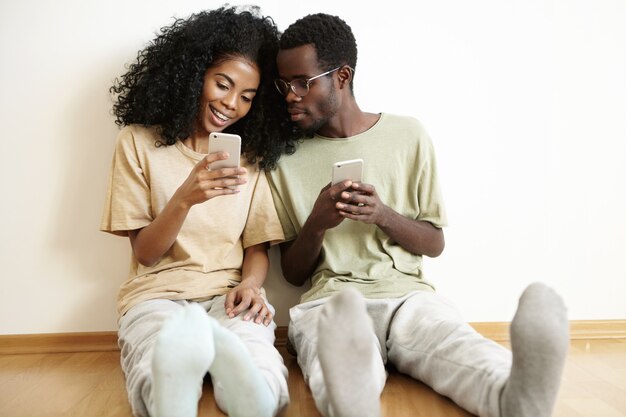 Cheerful young African woman with curly hair using smart phone, showing her pics on social networks to her handsome boyfriend, who is sitting next to her on floor